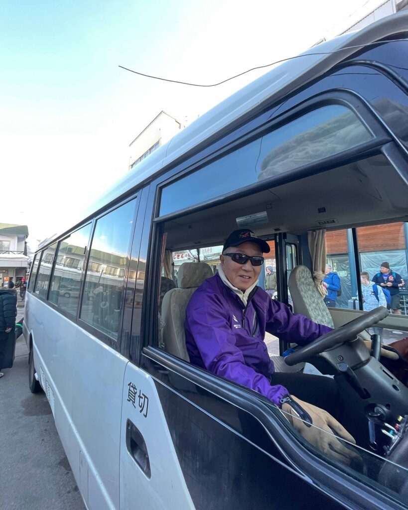 nagano snow shuttle bus driver wearing purple uniform jacket driving bus and smiling