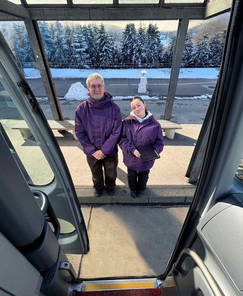 Two Nagano snow shuttle staff members wait to welcome customers off of a bus in the snowy Japanese landscape