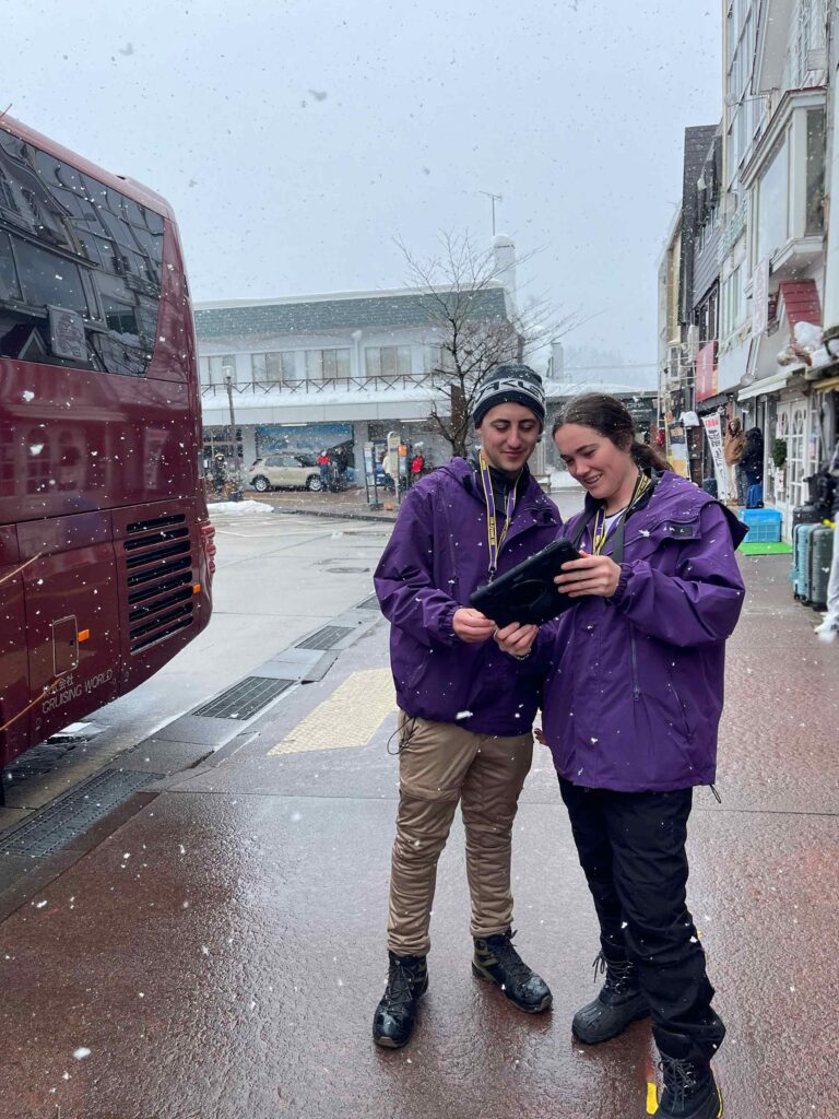 two nagano snow shuttle staff members wearing purple uniform jackets standing next to a bus and looking at a tablet