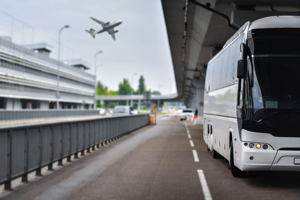 white shuttle bus witing at the airport for passengers a a plane flies overhead
